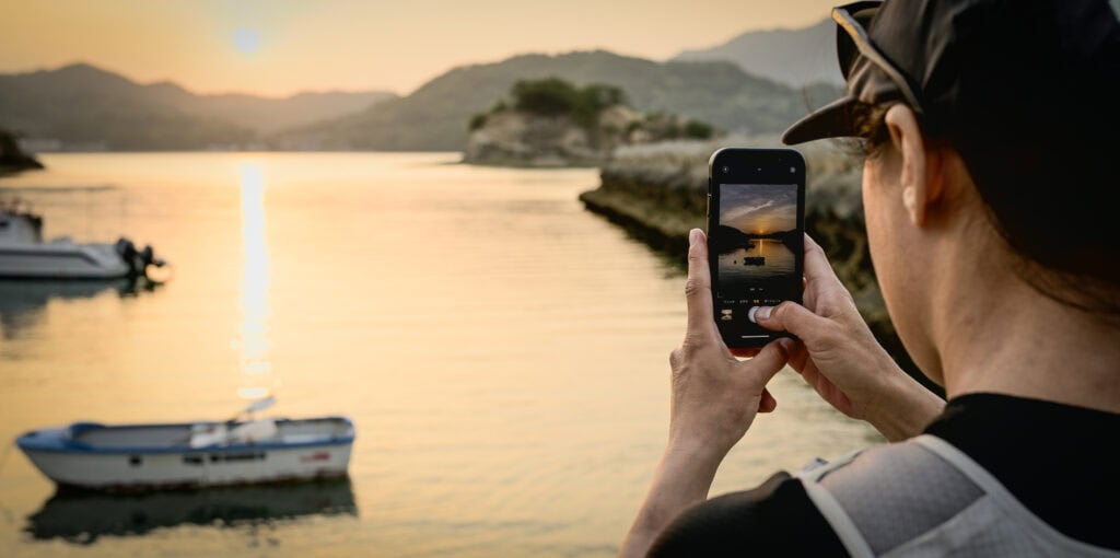Close-up of a cyclist taking a photo of a golden sunset over the water with a smartphone, small fishing boats and islands in the background