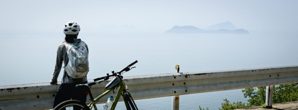 Cyclist with helmet and backpack resting beside a guardrail, gazing out over the calm Seto Inland Sea and misty islands in the distance