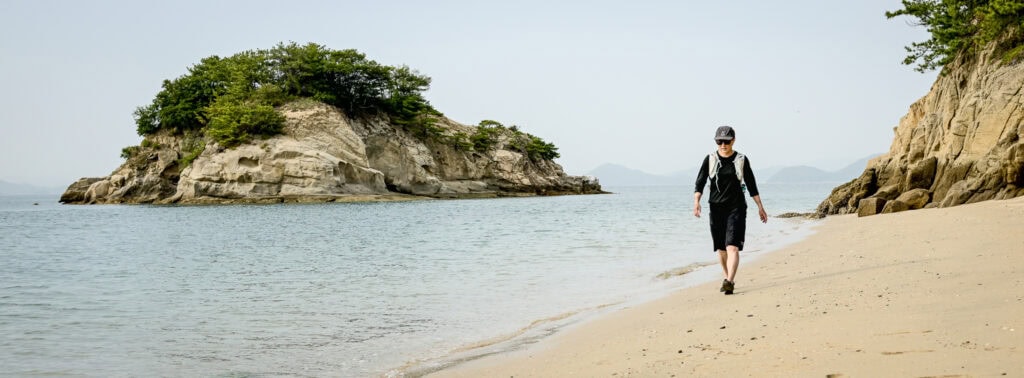 Person walking along a quiet sandy beach on Yumeshima Kaidō with rocky island cliffs in the background