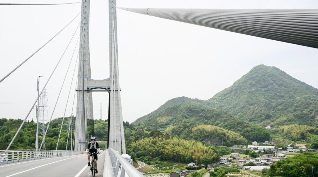 A cyclist rides across a suspension bridge on the Yumeshima Kaidō with green mountains and a rural Japanese village in the background.
