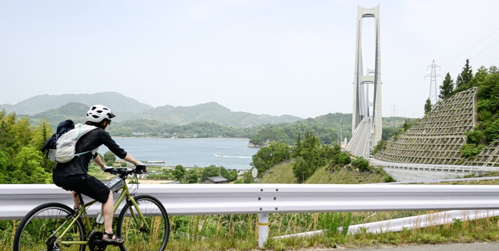 Cyclist overlooking the sea and bridge on Japan’s Yumeshima Kaidō, with mountains and water in the background.