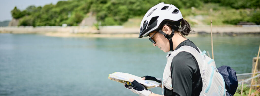 Woman cyclist wearing helmet and sunglasses reading a map near the water on Japan’s Yumeshima Kaidō cycling route.