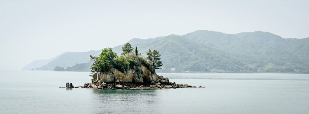 Small rocky island with pine trees surrounded by calm water in Japan’s Seto Inland Sea, with misty green mountains in the background.