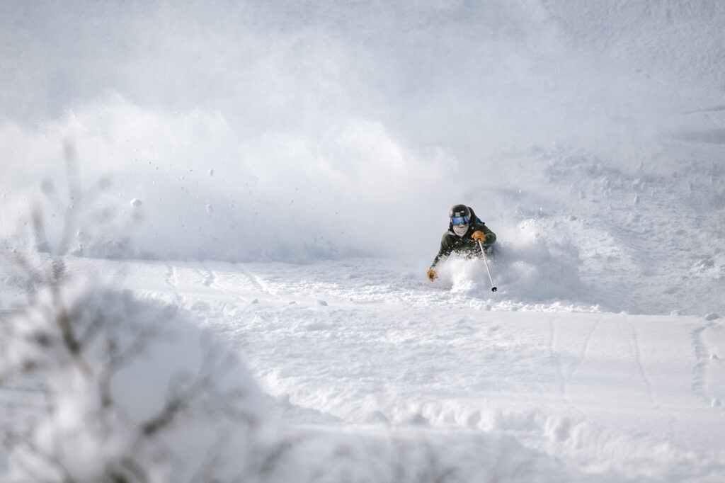Skiing through deep powder snow in Niseko, Japan, with a spray of snow exploding around them under clear winter light.