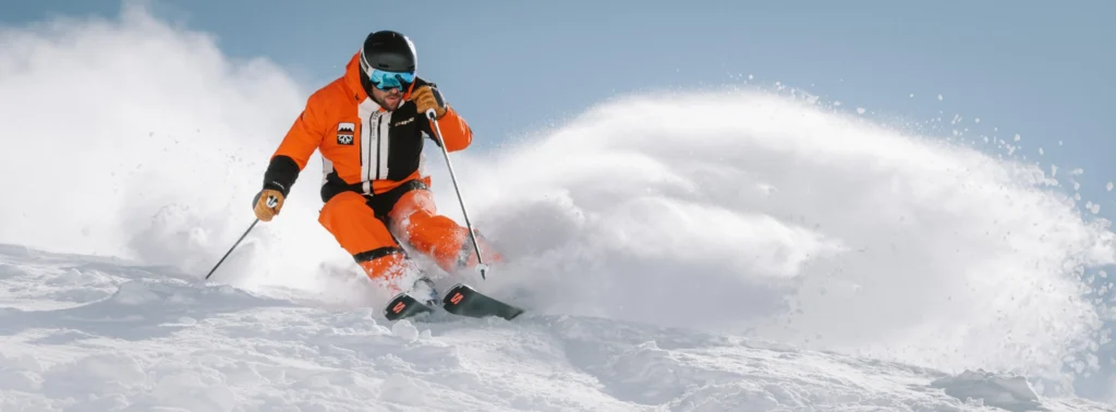Skier in orange jacket carving a fast turn with snow spray, demonstrating the value of reliable ski photography gear