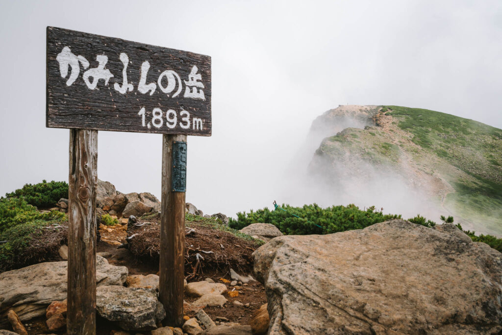 A weathered summit sign marking Kaminoyama (1,893m) in Daisetsuzan National Park, with mist drifting over the volcanic ridgeline near Tokachidake.