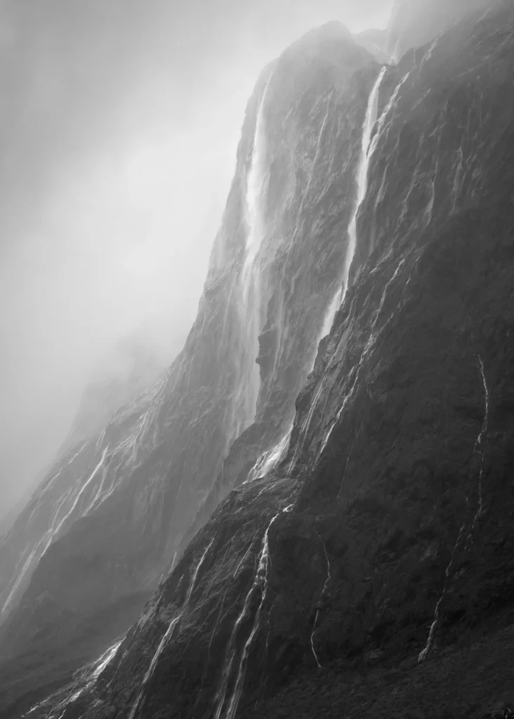 Black and white photo of misty waterfalls flowing down steep mountain cliffs in Fiordland, New Zealand
