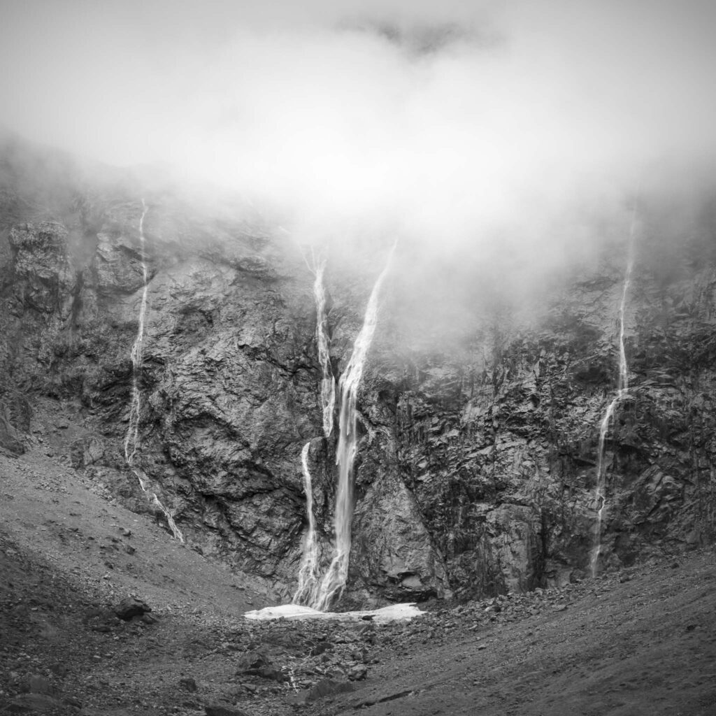 Black and white photo of thin waterfalls flowing down a rocky mountain face partly hidden in low cloud and mist in New Zealand